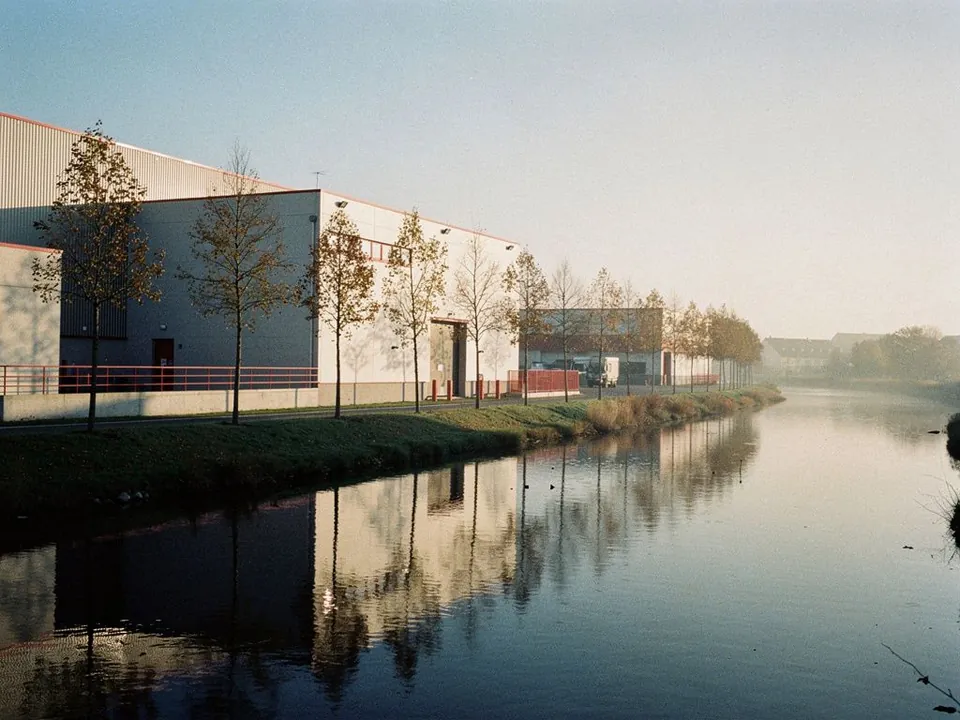 Factory beside a calm river lined with young trees in morning light
